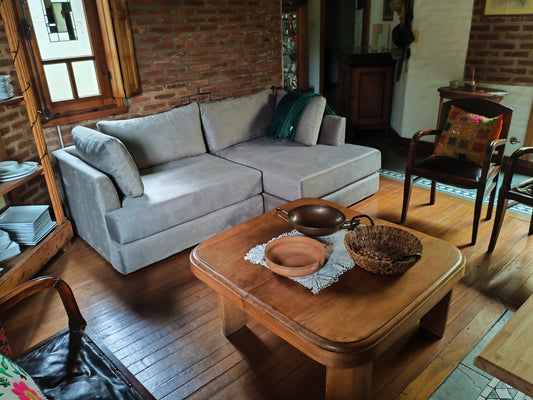 Sillón esquinero tapizado en tela gris claro, de líneas rectas y diseño moderno, acompañado de almohadones al tono y ubicado en un ambiente rústico con paredes de ladrillo visto y piso de madera.