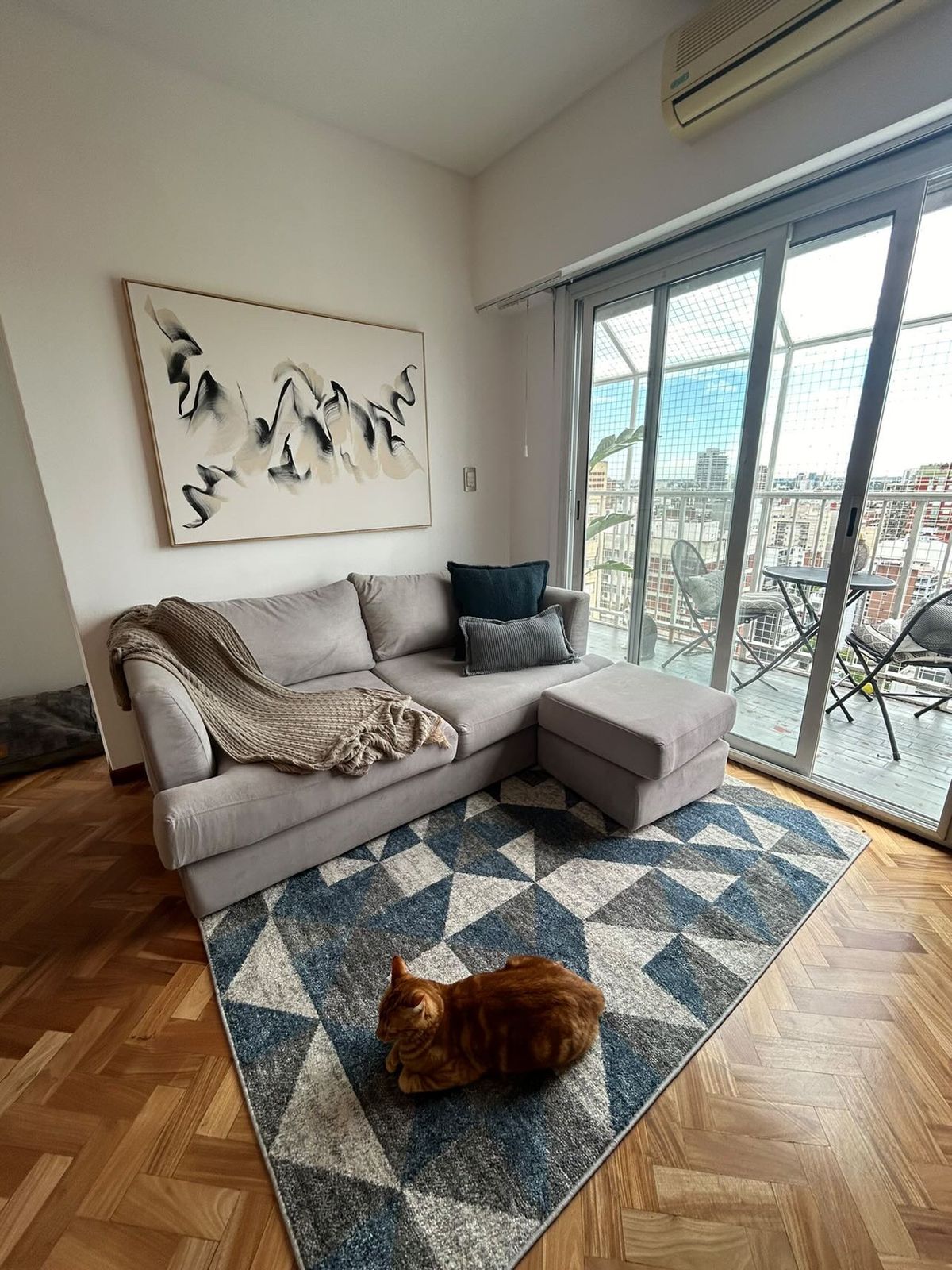 Sillón esquinero tapizado en tono gris claro, con almohadones decorativos en azul y gris, acompañado de una mesa auxiliar de madera sobre una alfombra geométrica en tonos azules y blancos, en un living luminoso y moderno