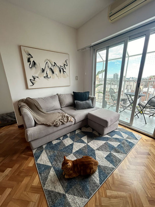 Sillón esquinero tapizado en tono gris claro, con almohadones decorativos en azul y gris, acompañado de una mesa auxiliar de madera sobre una alfombra geométrica en tonos azules y blancos, en un living luminoso y moderno