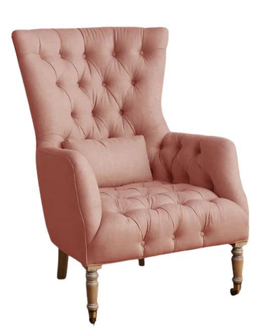 Sillón individual tapizado en rosa claro, con respaldo alto y capitoné, cojín lumbar suelto, brazos curvos y patas delanteras torneadas en madera clara.