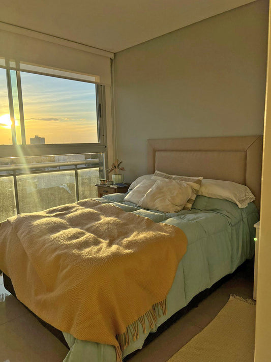 Respaldo de cama tapizado en tela color beige claro, de diseño recto y minimalista, acompañando una cama con acolchado celeste y almohadones decorativos en tonos neutros.