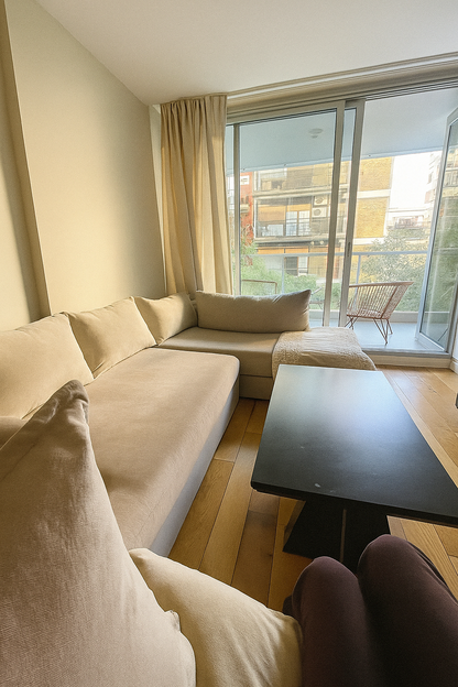 Living luminoso con mesa ratona rectangular negra, sofá esquinero beige y sillón de un cuerpo a juego, frente a un ventanal corredizo.