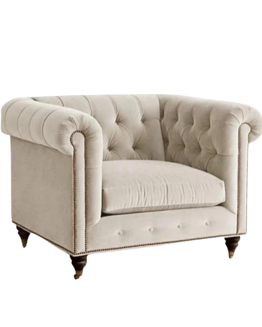 Sillón individual estilo Chesterfield tapizado en color gris claro, con capitoné en respaldo y apoyabrazos, tachas decorativas y patas de madera torneadas.