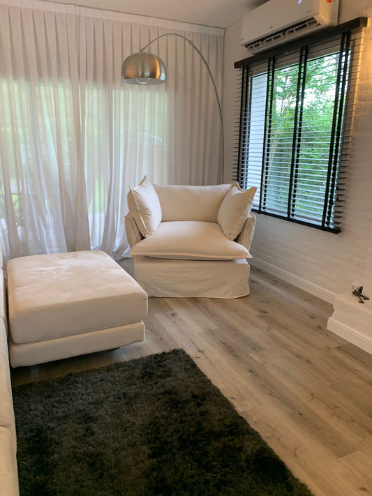 Sillón individual tapizado en color beige claro, con almohadones amplios y mullidos, ubicado en un ambiente luminoso junto a una ventana con cortinas y piso de madera clara.