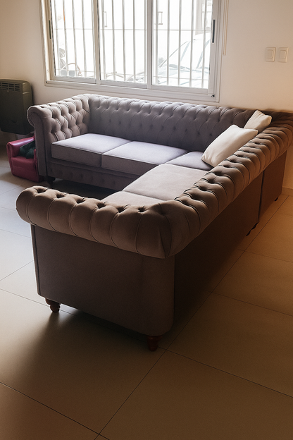 Sillón esquinero estilo Chesterfield en tapizado de terciopelo color gris topo, con respaldo y apoyabrazos capitoné, cojines lisos y patas de madera