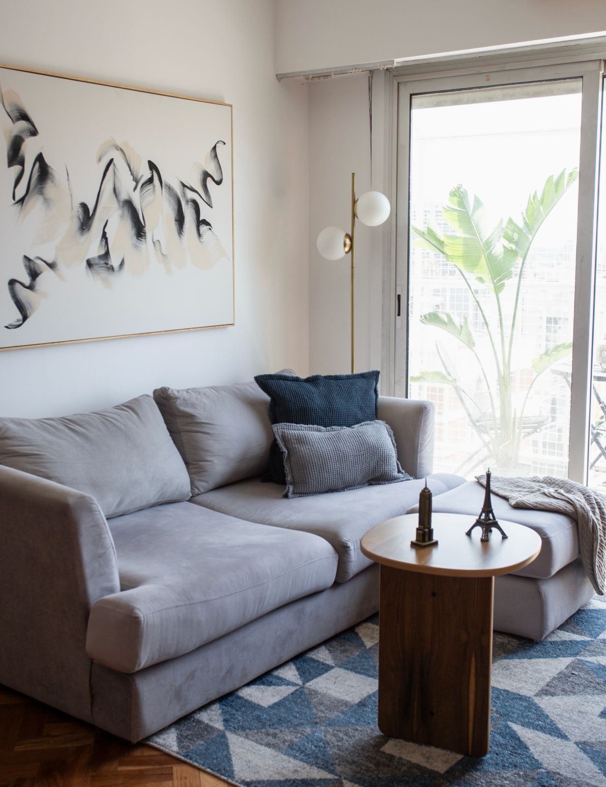 Sillón esquinero tapizado en tono gris claro, con almohadones decorativos en azul y gris, acompañado de una mesa auxiliar de madera sobre una alfombra geométrica en tonos azules y blancos, en un living luminoso y moderno
