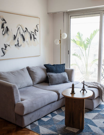 Sillón esquinero tapizado en tono gris claro, con almohadones decorativos en azul y gris, acompañado de una mesa auxiliar de madera sobre una alfombra geométrica en tonos azules y blancos, en un living luminoso y moderno