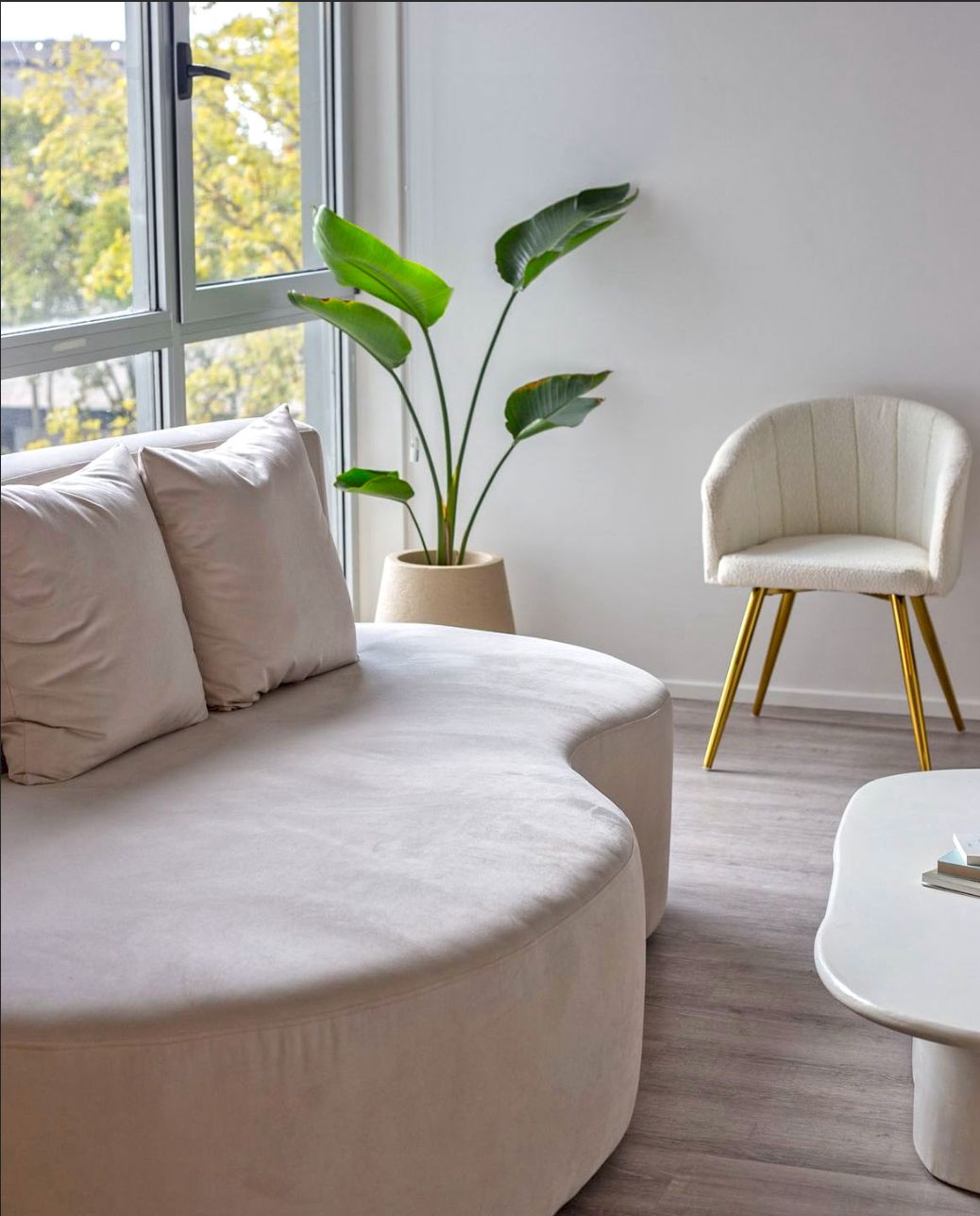 Sillón moderno de forma curva, tapizado en tono claro, con tres cojines decorativos, ubicado junto a una ventana grande que permite la entrada de luz natural, acompañado de plantas en macetas que aportan frescura al espacio