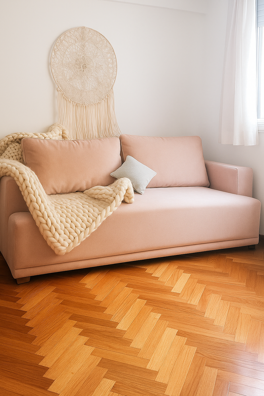 Sillón de dos cuerpos en color rosa viejo con líneas rectas y apoyabrazos anchos, decorado con un cojín gris claro y una manta de lana gruesa en tono beige. Sobre la pared blanca cuelga un atrapasueños artesanal y el ambiente se ilumina con luz natural que entra por la ventana, destacando el piso de parquet en diseño espiga