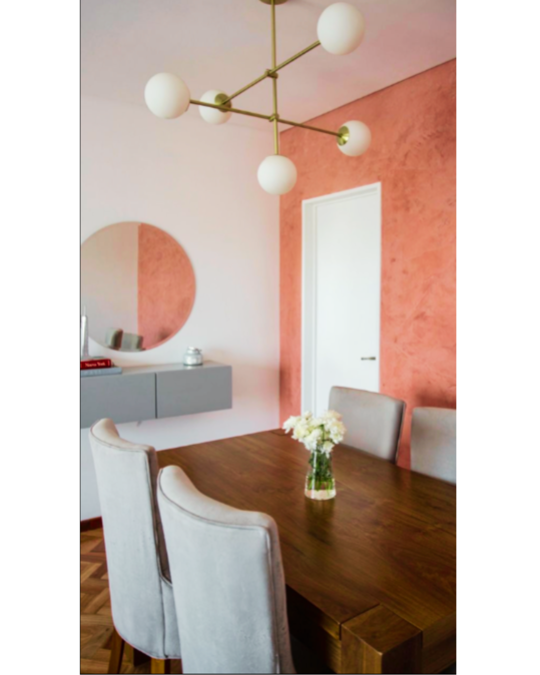 Comedor contemporáneo con pared de acento rosa, mesa de madera maciza, sillas tapizadas en gris claro y lámpara colgante moderna de esferas blancas. En la pared hay un espejo redondo sobre un mueble flotante gris.