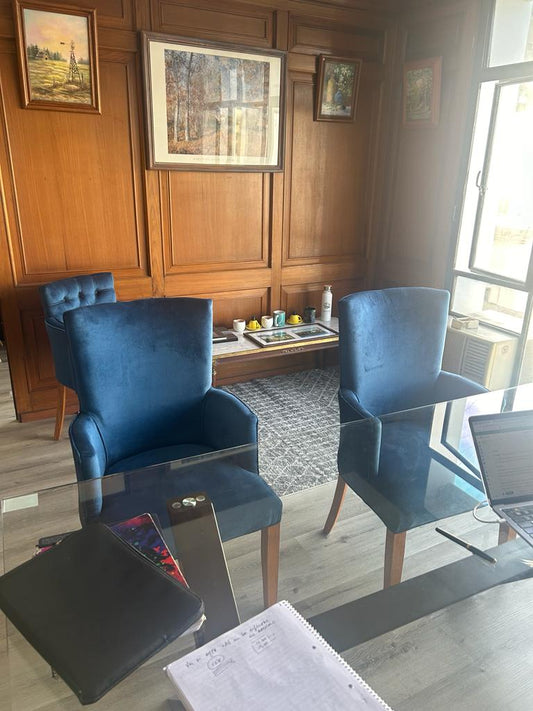 Sillas de comedor tapizadas en terciopelo azul con patas de madera, ubicadas junto a una mesa de vidrio en un ambiente elegante con paredes de madera.
