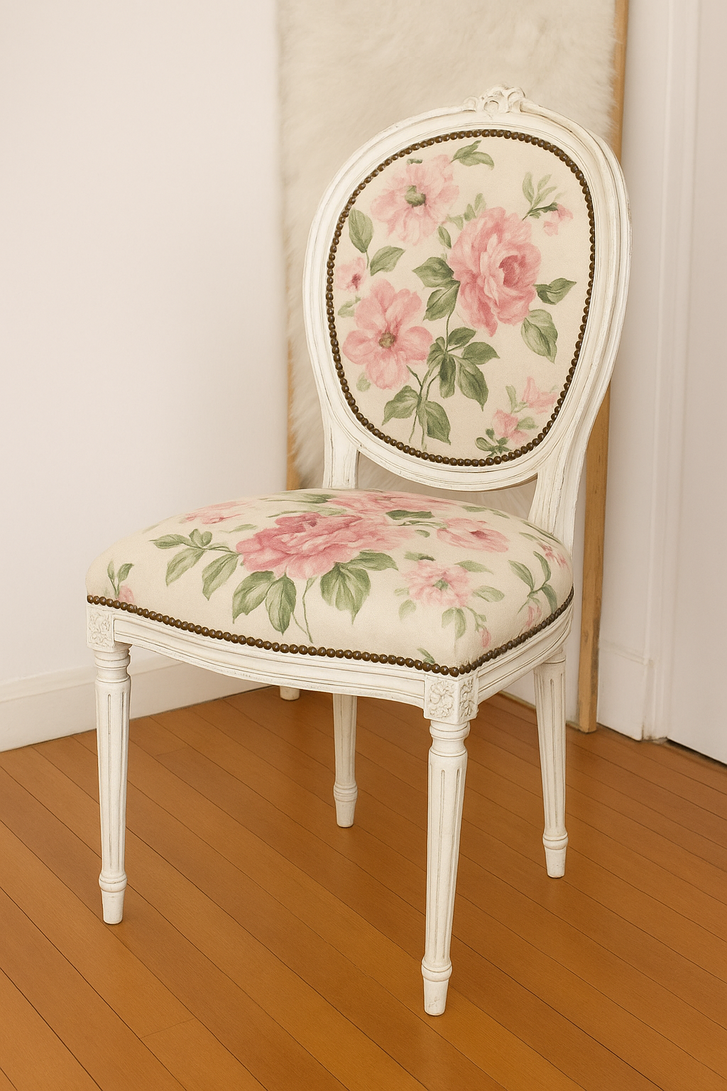 Silla de estilo vintage con estructura de madera tallada pintada en blanco envejecido. El respaldo ovalado y el asiento están tapizados con tela de fondo claro y estampado floral de grandes rosas rosadas y hojas verdes. Las patas son torneadas y decoradas con detalles clásicos, evocando un diseño francés o victoriano. La silla está colocada sobre un suelo de madera clara en un rincón luminoso.
