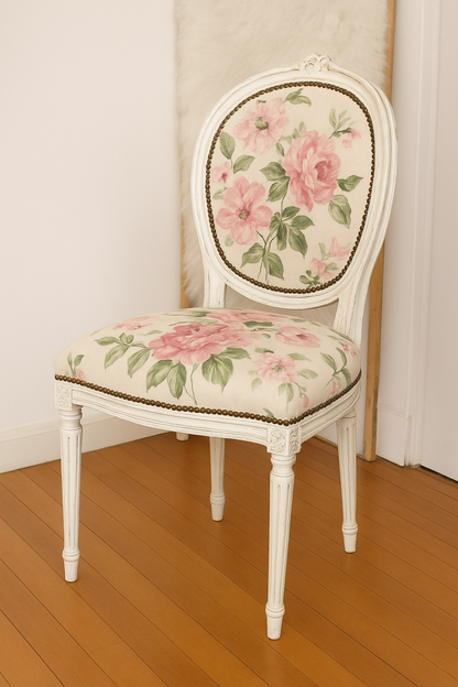 Silla de estilo vintage con estructura de madera tallada pintada en blanco envejecido. El respaldo ovalado y el asiento están tapizados con tela de fondo claro y estampado floral de grandes rosas rosadas y hojas verdes. Las patas son torneadas y decoradas con detalles clásicos, evocando un diseño francés o victoriano. La silla está colocada sobre un suelo de madera clara en un rincón luminoso.