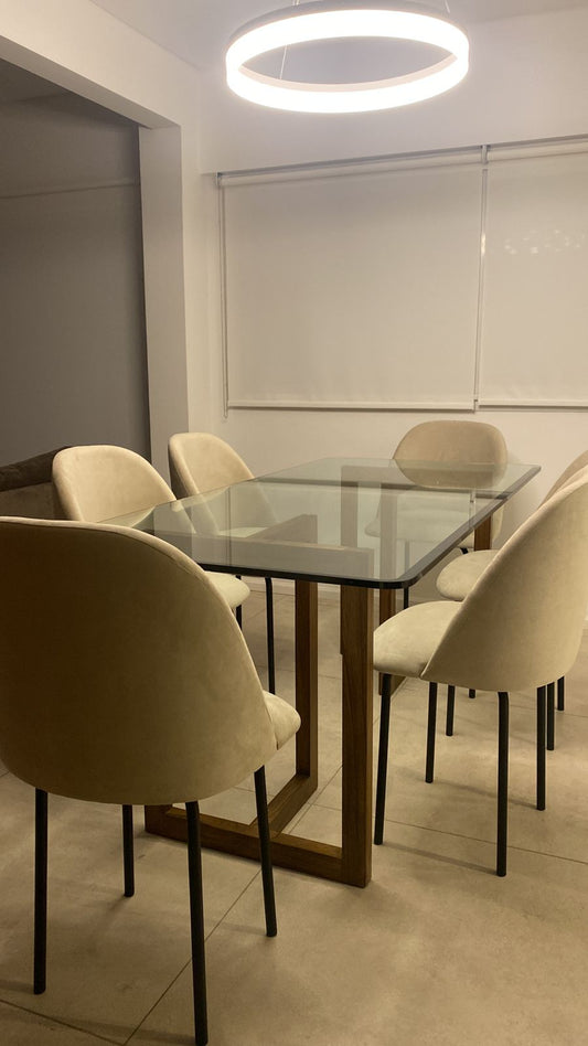 Sillas de comedor tapizadas en tela beige claro, con respaldo curvo y patas delgadas de metal negro, dispuestas alrededor de una mesa rectangular de vidrio con base de madera