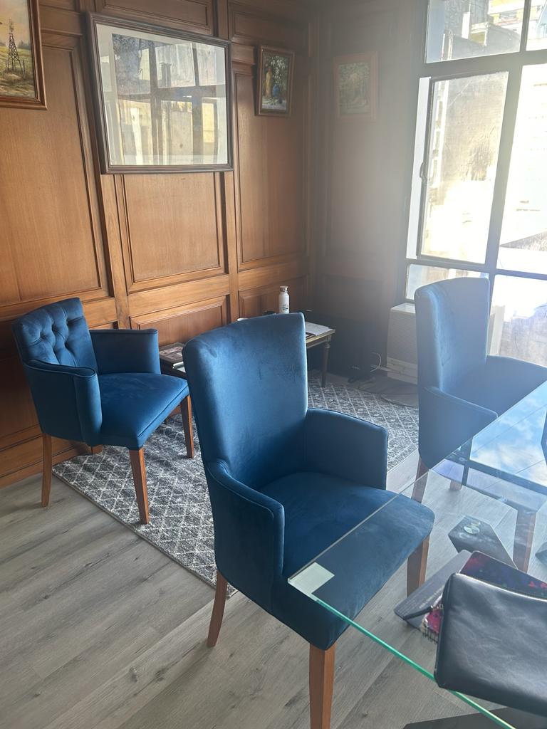 Sillas de comedor tapizadas en terciopelo azul con patas de madera, ubicadas junto a una mesa de vidrio en un ambiente elegante con paredes de madera.