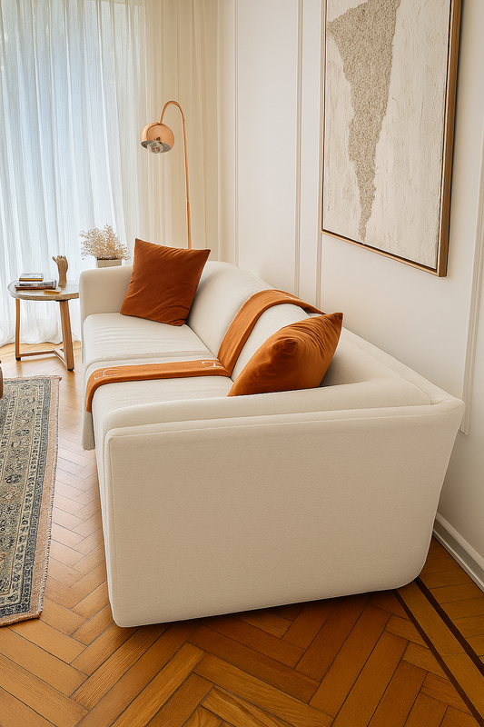 Sillón de tres cuerpos color blanco con dos almohadones de terciopelo terracota y una manta en tonos naranjas, ubicado en un living luminoso con mesa ratona de vidrio y alfombra persa.