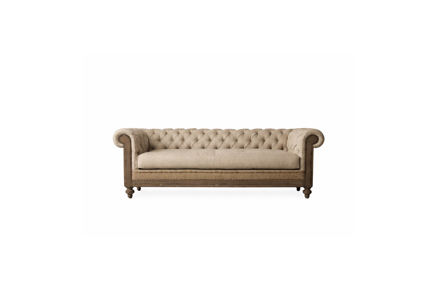 Fotografía editorial de interiores que muestra un living sofisticado y tradicional. El protagonista es un sillón Chesterfield de tres cuerpos, tapizado en lino texturizado color beige, con capitoné profundo, brazos enrollados y una estructura de madera envejecida con detalles de tachuelas. El sillón se apoya sobre una alfombra persa antigua sobre piso de madera. Al fondo, una gran biblioteca empotrada de piso a techo, hecha de madera oscura, está llena de libros y objetos decorativos como jarrones de latón 