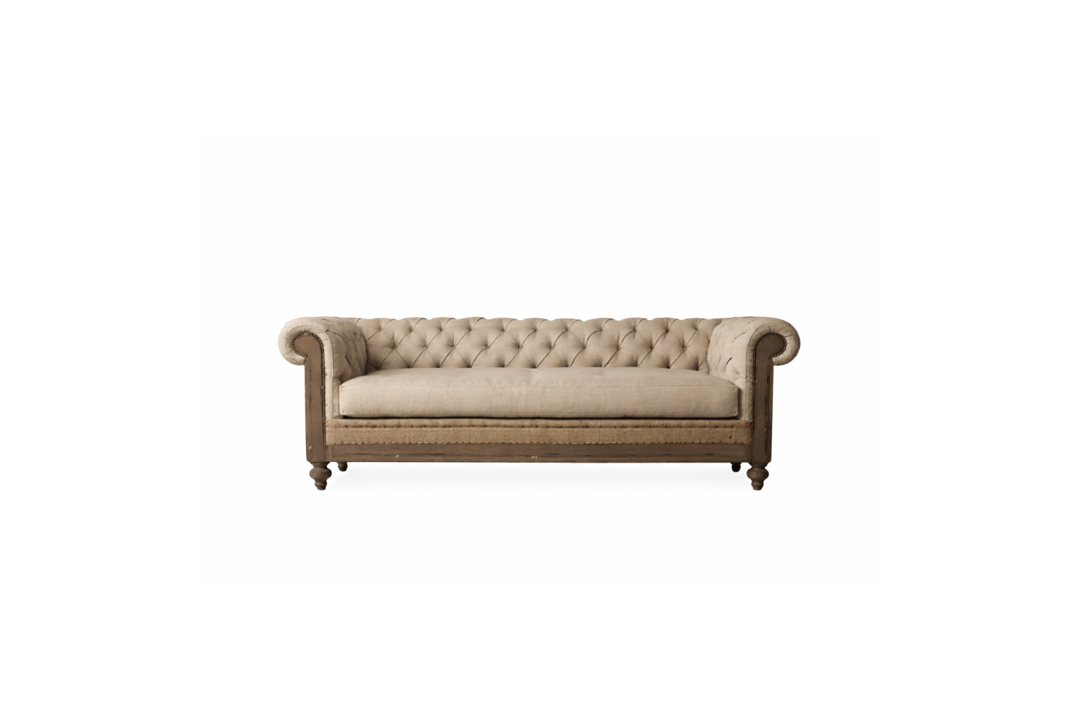 Fotografía editorial de interiores que muestra un living sofisticado y tradicional. El protagonista es un sillón Chesterfield de tres cuerpos, tapizado en lino texturizado color beige, con capitoné profundo, brazos enrollados y una estructura de madera envejecida con detalles de tachuelas. El sillón se apoya sobre una alfombra persa antigua sobre piso de madera. Al fondo, una gran biblioteca empotrada de piso a techo, hecha de madera oscura, está llena de libros y objetos decorativos como jarrones de latón 