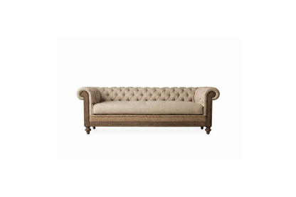 Fotografía editorial de interiores que muestra un living sofisticado y tradicional. El protagonista es un sillón Chesterfield de tres cuerpos, tapizado en lino texturizado color beige, con capitoné profundo, brazos enrollados y una estructura de madera envejecida con detalles de tachuelas. El sillón se apoya sobre una alfombra persa antigua sobre piso de madera. Al fondo, una gran biblioteca empotrada de piso a techo, hecha de madera oscura, está llena de libros y objetos decorativos como jarrones de latón 