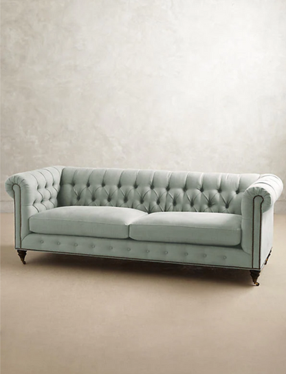 Sofá clásico Chesterfield, tapizado en pana, con respaldo y apoyabrazos capitoné, líneas elegantes y sofisticadas, patas de madera oscura tallada, sobre fondo blanco, ideal para decorar livings, oficinas o salas de estilo tradicional con un toque de lujo.