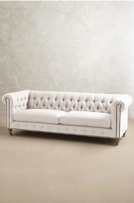 Sofá clásico Chesterfield, tapizado en pana, con respaldo y apoyabrazos capitoné, líneas elegantes y sofisticadas, patas de madera oscura tallada, sobre fondo blanco, ideal para decorar livings, oficinas o salas de estilo tradicional con un toque de lujo.