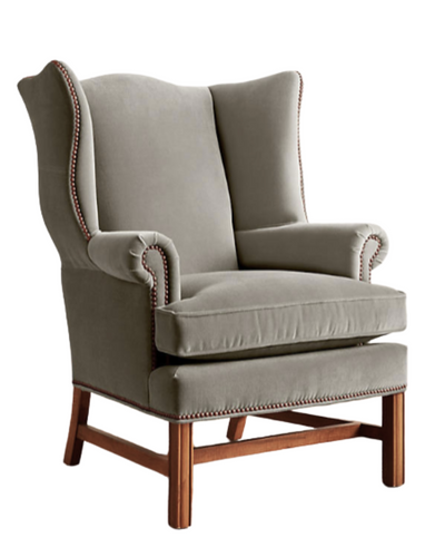 Sillón individual tapizado en terciopelo gris claro, con respaldo alto y orejero, cojines sueltos en asiento y respaldo, y base de madera vista con patas cruzadas.