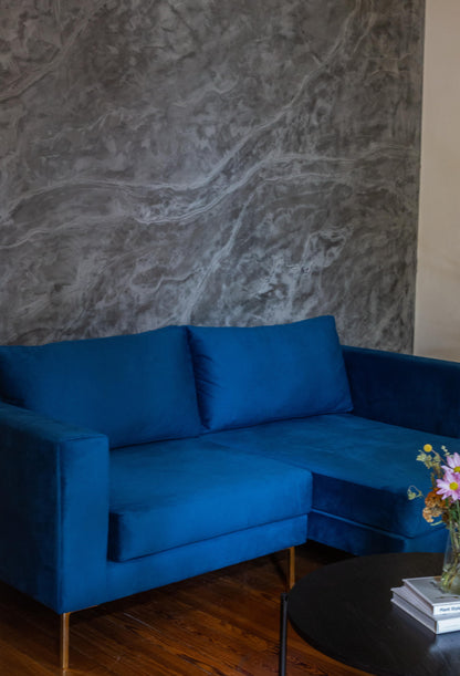 Sillón esquinero moderno en azul con patas doradas, líneas limpias y almohadones sueltos. Estilo nórdico con toque glam.