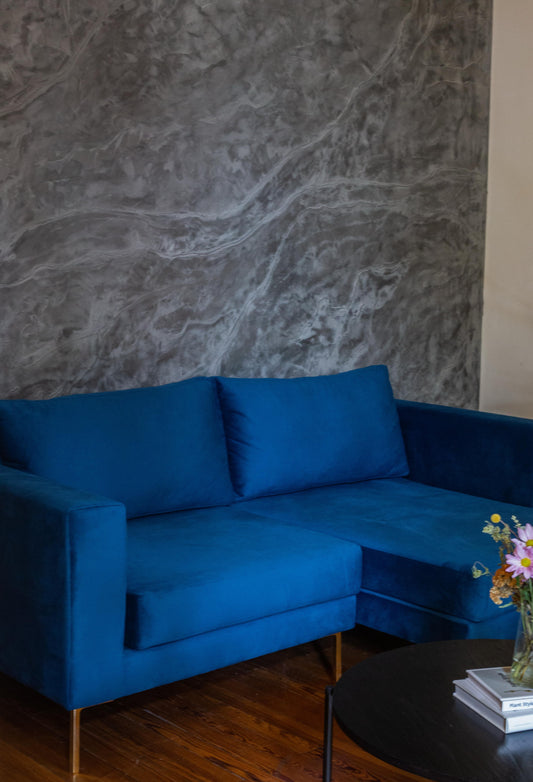 Sillón esquinero moderno en azul con patas doradas, líneas limpias y almohadones sueltos. Estilo nórdico con toque glam.