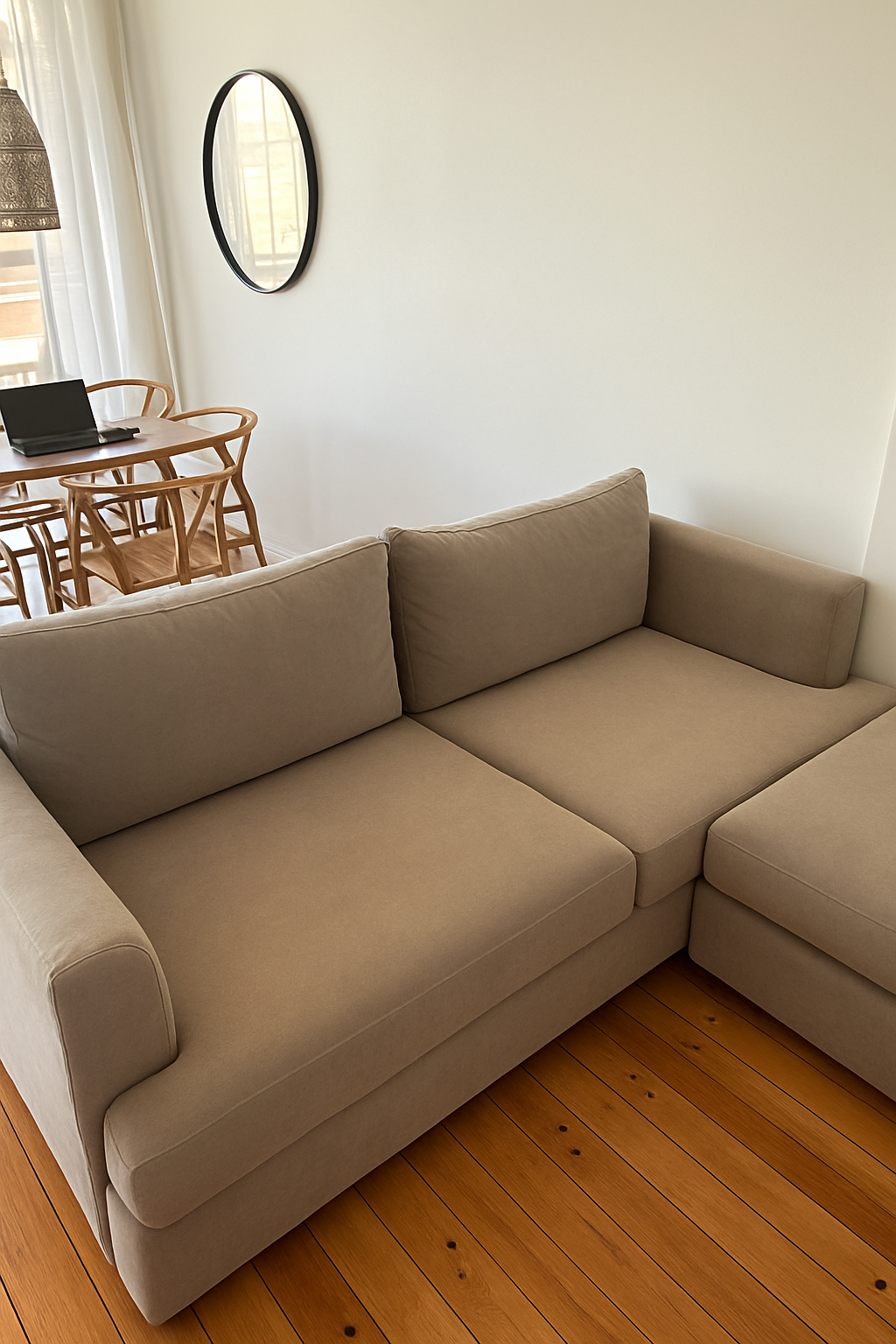 Sillón esquinero tapizado en tono beige, de líneas rectas y cojines amplios, ubicado sobre un piso de madera lustrada junto a una mesa ratona redonda de mármol.