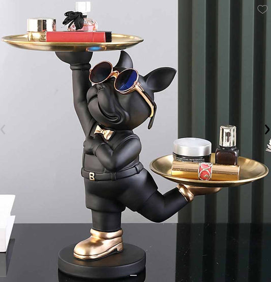 Bulldog francés con traje negro y zapatos dorados que sostiene dos bandejas doradas con perfumes y crema