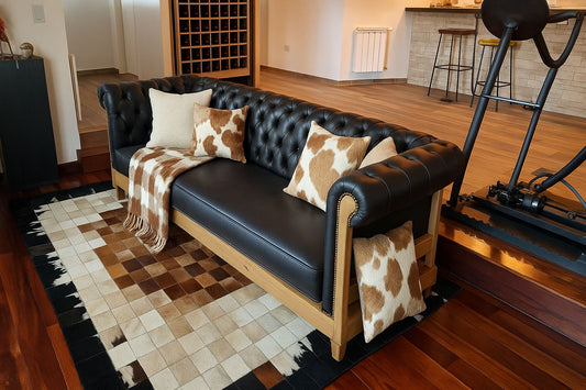 Sofá tipo Chesterfield de tres plazas con estructura de madera clara expuesta y tapizado en cuero negro, con característicos brazos y respaldo acolchados en capitoné. Está decorado con cojines en tonos beige y estampado de piel de vaca, junto con una manta a juego que cuelga sobre uno de los brazos