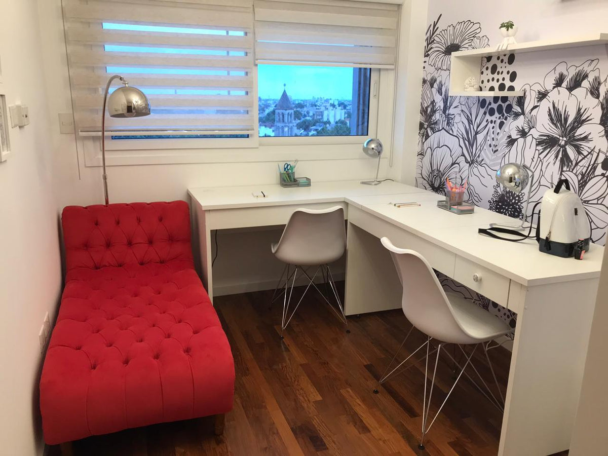 Diván Chesterfield rojo con capitoné profundo, sin brazos, junto a un escritorio blanco en L y un papel floral, en el consultorio de Agustina