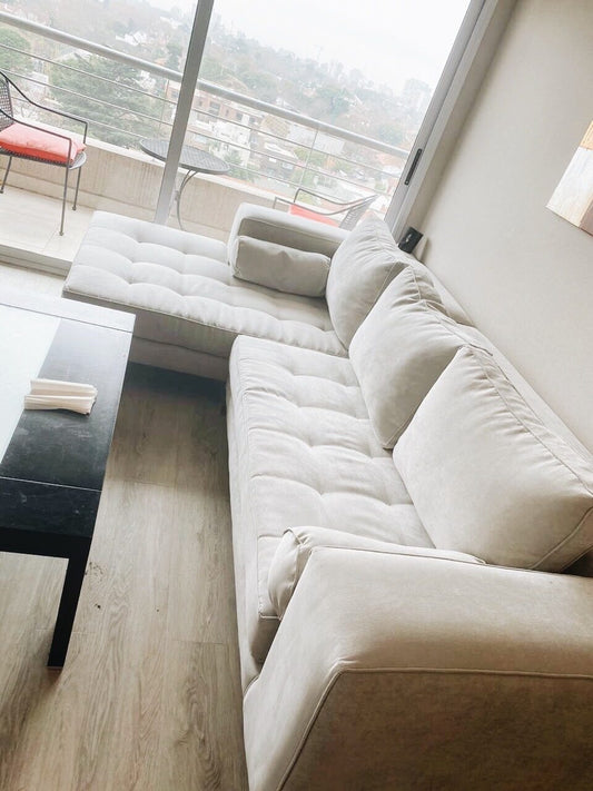 Esquinero Bambú beige con asiento capitoné y almohadón cilíndrico, frente al balcón con vista a la ciudad de Belén