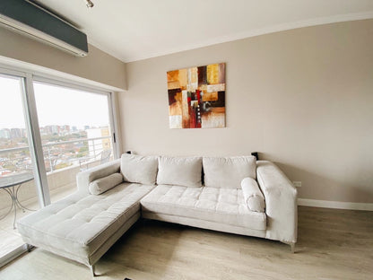 Esquinero Bambú beige con asiento capitoné y almohadones cilíndricos, junto al balcón con vista a la ciudad en el living de Brenda