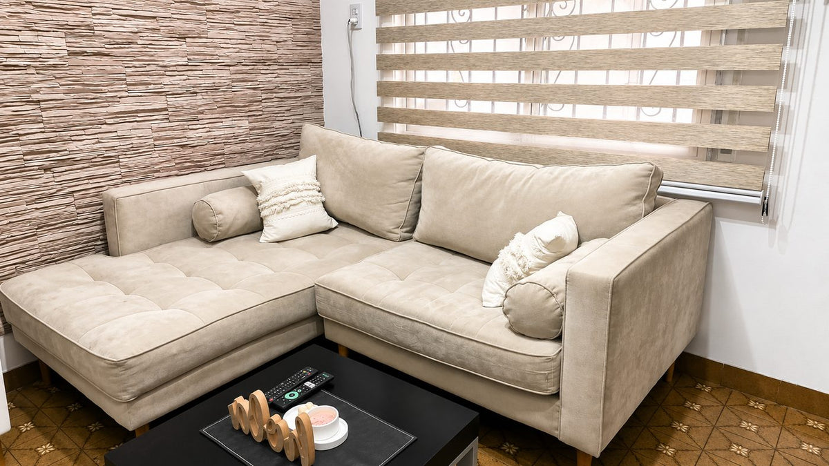 Esquinero Bambú beige claro con asiento capitoné y almohadones cilíndricos, junto a la pared de piedra del living de Rocío
