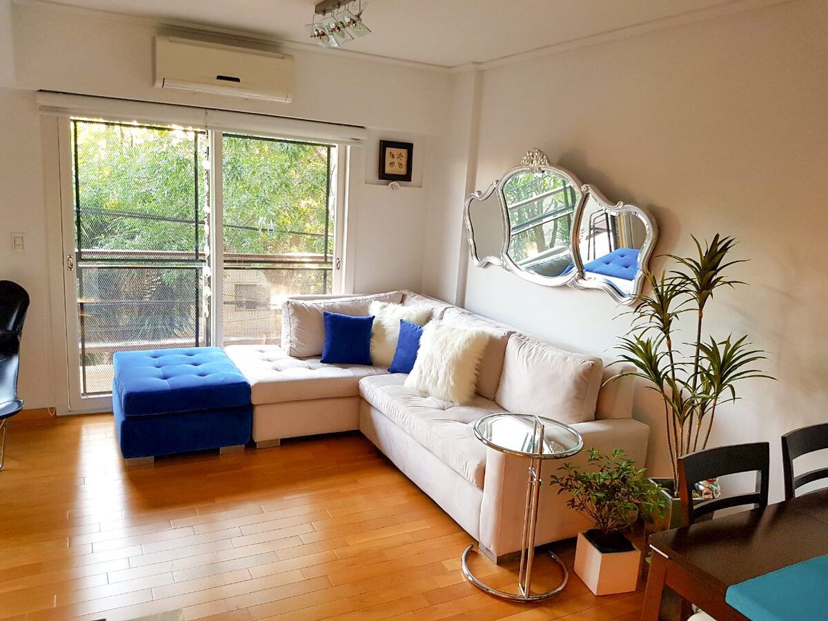 Sillón esquinero crema con asiento capitoné y puff azul eléctrico, frente a un espejo barroco plateado, en el living de Nerina