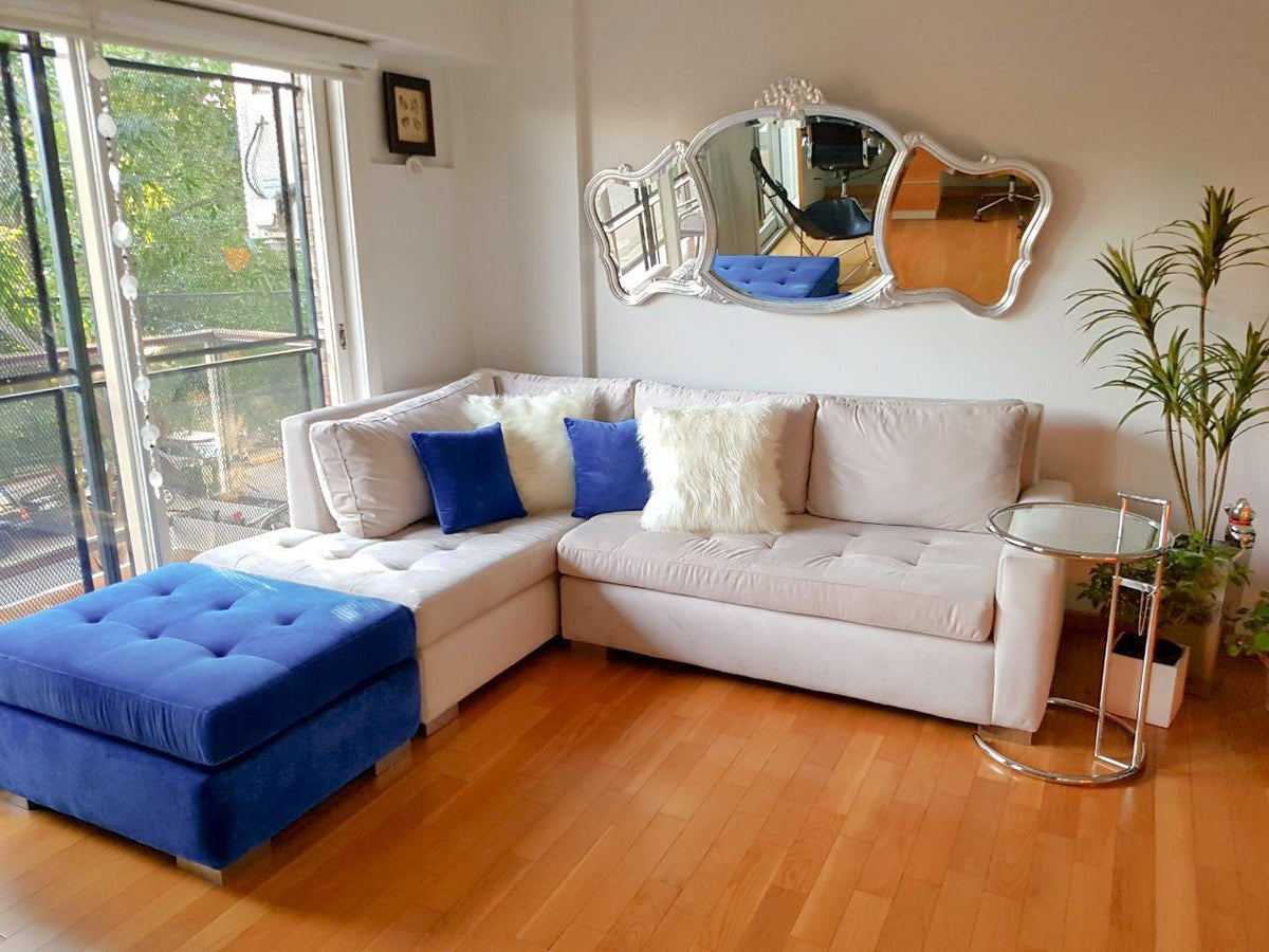 Sillón esquinero en tono crudo con asiento capitoné y puff azul capitoné en un living con balcón y espejo
