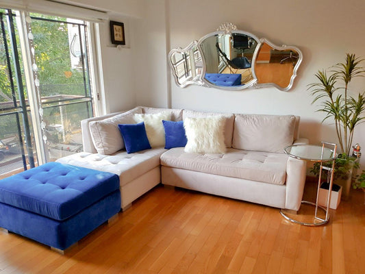 Sillón esquinero en tono crudo con asiento capitoné y puff azul capitoné en un living con balcón y espejo