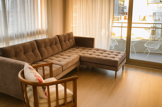 Sofá esquinero tapizado en tela marrón claro con diseño capitoné en asiento y respaldo. Tiene patas de madera delgadas y está ubicado en una sala iluminada por luz natural que entra por grandes ventanales con cortinas translúcidas