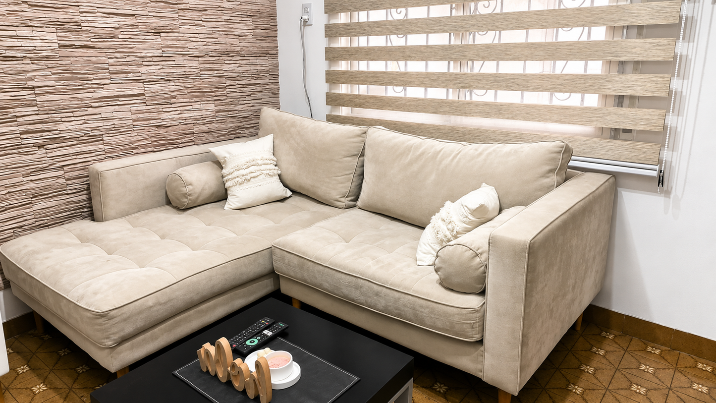 Fotografía profesional de un living moderno y acogedor. Sofá esquinero en tono beige claro con tapizado aterciopelado, almohadones decorativos y patas de madera natural. Pared revestida con piedra texturada color arena, cortinas roller dúo beige y mesa de centro negra minimalista. Iluminación cálida y uniforme, imagen nítida en alta resolución, colores equilibrados, sombras suaves, ambiente elegante y luminoso. Perspectiva amplia, estilo de revista de decoración, materiales realistas, acabados premium, máx