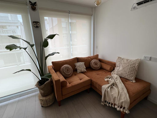 Un sillón esquinero tapizado en pana color terracota, decorado con almohadones tejidos en tonos neutros y una manta clara, que crean un ambiente cálido y natural en el living