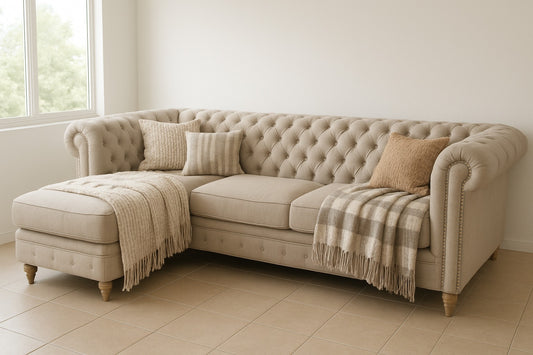 fá esquinero estilo Chesterfield tapizado en tela beige claro, con diseño capitoné en el respaldo y brazos enrollados. El sofá tiene patas de madera torneadas en tono natural, y está decorado con cojines en tonos tierra y mantas tejidas en colores neutros, que cuelgan suavemente sobre los brazos.
