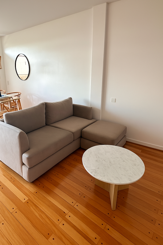 Sillón esquinero tapizado en tono beige, de líneas rectas y cojines amplios, ubicado sobre un piso de madera lustrada junto a una mesa ratona redonda de mármol.