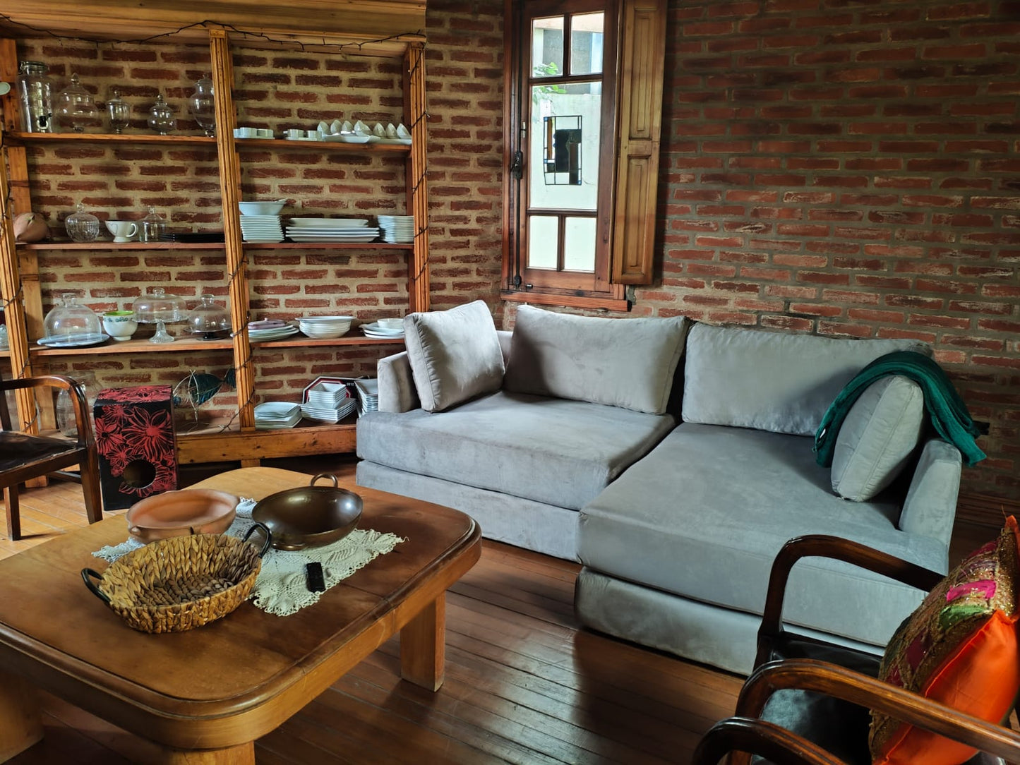 Sillón esquinero tapizado en tela gris claro, de líneas rectas y diseño moderno, acompañado de almohadones al tono y ubicado en un ambiente rústico con paredes de ladrillo visto y piso de madera.