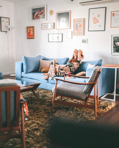 Living de Josefina Álvarez Castillo con su Sofá Elton azul, una butaca de madera y una pared de cuadros