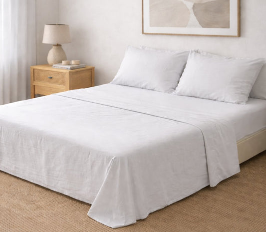 Juego de sábanas lisas Queen blanco con dos fundas de almohada, en cama grande junto a una mesa de luz