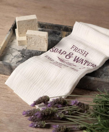 Toalla de mano blanca con leyenda Fresh Soap and Water junto a jabones y ramitas de lavanda