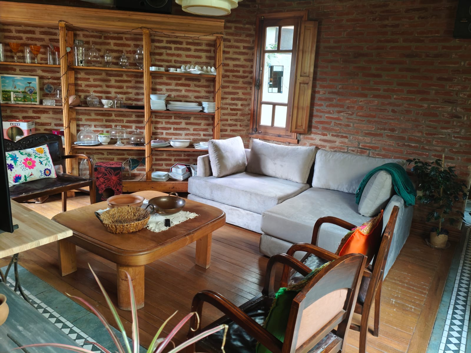 Sillón esquinero tapizado en tela gris claro, de líneas rectas y diseño moderno, acompañado de almohadones al tono y ubicado en un ambiente rústico con paredes de ladrillo visto y piso de madera.