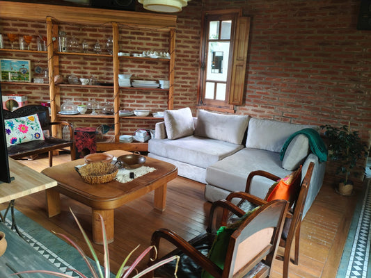 Sillón esquinero tapizado en tela gris claro, de líneas rectas y diseño moderno, acompañado de almohadones al tono y ubicado en un ambiente rústico con paredes de ladrillo visto y piso de madera.