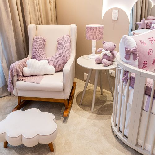 Mecedora Ciruelo en crema con almohada de lactancia lila, manta rosa y almohadón nube, junto a un puff blanco y una cuna redonda